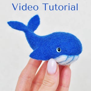 May include: A blue felt whale with a white belly and black eyes. The whale is being held in a hand.