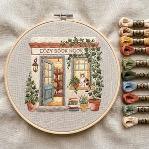 Puede incluir: Un bastidor de bordado con un panel de tela que muestra una escena bordada de una librería con la etiqueta COZY BOOK NOOK. El diseño incluye un gato calicó en una ventana, plantas en macetas y una pila de libros. Diez madejas de hilo están alineadas.