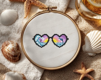 May include: A cross-stitch embroidery hoop featuring heart-shaped sunglasses with a beach scene. The sunglasses have a black outline and are filled with pixelated images of waves, dolphins, and a sunset. The hoop is set on a sandy surface with seashells and starfish.