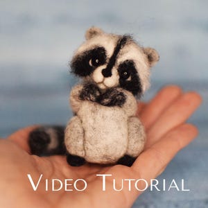 Peut inclure: Un petit raton laveur en feutre aiguille, gris et noir, avec un nez noir et des yeux noirs. Le raton laveur est assis sur une main, les pattes croisées devant lui. L'arrière-plan est une surface bleu clair. L'image comporte le texte "VIDEO TUTORIAL" en bas.