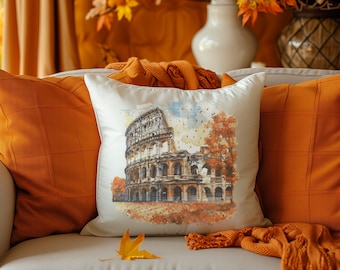 May include: A square cushion with a watercolour image of the Colosseum in Rome, Italy, set against autumnal trees. The cushion sits on a cream sofa, with orange cushions and a knitted throw. The scene evokes a warm, autumnal atmosphere.