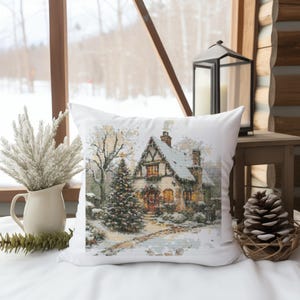 Könnte beinhalten: Ein weißes Kissen mit einer Winterszene eines schneebedeckten Häuschens mit einem beleuchteten Fenster und einem Weihnachtsbaum im Vorgarten. Das Häuschen hat eine rote Tür und ein schneebedecktes Dach.