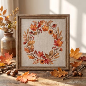 May include: A square wooden frame containing a cross-stitch embroidery of an autumn wreath. The wreath features orange, red, and yellow maple leaves, wheat stalks, small berries, and flowers arranged in a circle on a light beige fabric background.