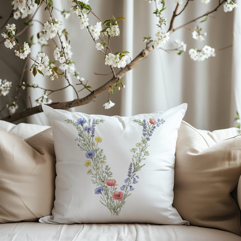 May include: White square pillow with a floral "V" design in blue, yellow, and red. The pillow is on a beige sofa, with a flowering tree branch in the background.