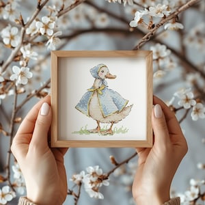 May include: A framed cross-stitch of a duck wearing a blue cape and bonnet with a yellow bow. The duck stands on green grass. The frame is made of light brown wood. The background features white flowers and branches.