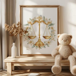May include: A framed print featuring a golden scale of justice surrounded by a floral wreath. The artwork is set against a white background and framed in a light brown wooden frame. A beige teddy bear sits on a wooden bench.