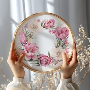 Puede incluir: Un aro de bordado dorado con fondo blanco y un diseño floral. El diseño presenta flores rosas y blancas, hojas y tallos.