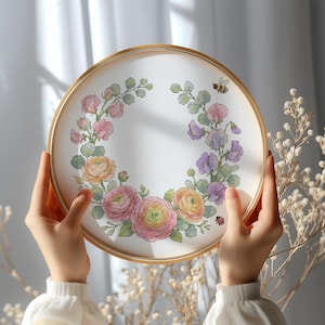 Könnte beinhalten: Ein runder Stickrahmen mit einem floralen Kreuzstichmuster. Das Design zeigt rosa, gelbe und lila Blumen, grüne Blätter, eine Biene und einen Marienkäfer. Der Rahmen hat einen goldfarbenen Holzrahmen.