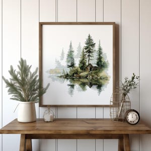 Puede incluir: Una impresión en acuarela enmarcada representa una serena escena junto al lago con una pequeña cabaña entre árboles de hoja perenne. La obra de arte se exhibe sobre una mesa de madera, junto con elementos decorativos como un jarrón con vegetación, una vela y una jaula de alambre con un reloj.