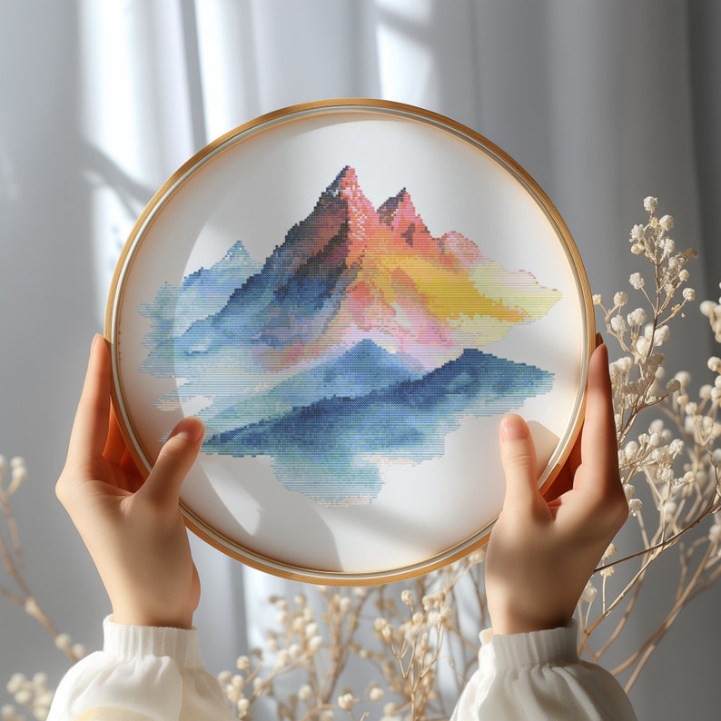 May include: A framed cross-stitch embroidery depicting a colorful mountain range with a watercolor effect. The mountains are shades of pink, orange, yellow, blue, and green. The embroidery is in a round, wooden frame.
