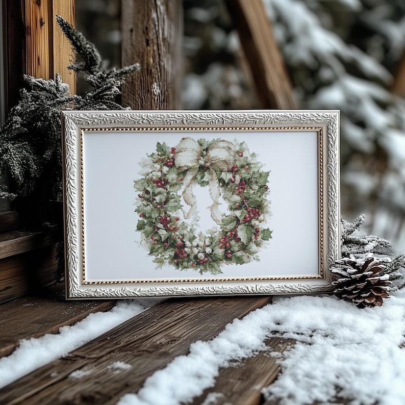 May include: A framed print of a Christmas wreath with a cream-colored bow. The wreath features green leaves, red berries, and white accents. The frame is white with a gold inner border. The image is set on a wooden surface with snow.