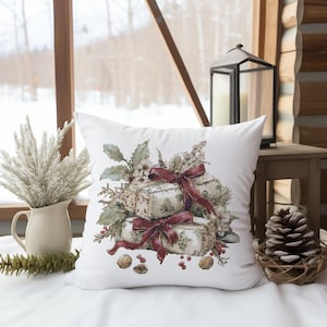 May include: White square decorative pillow featuring a watercolor illustration of wrapped gifts with red ribbons and greenery. The pillow is decorated with a floral design and is placed on a white surface.
