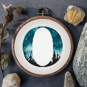 Peut inclure: Une broderie au point de croix dans un cercle en bois présente un motif de ciel nocturne bleu sarcelle et blanc avec un grand "O" blanc. Le motif comprend des silhouettes d'arbres en bas et des étoiles. Le cercle est posé sur une surface gris foncé.