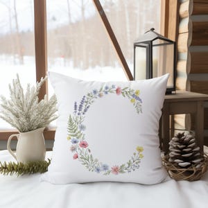 May include: White square pillow with a floral wreath in the shape of the letter "C". The wreath features a variety of colorful flowers and greenery. The pillow is on a white surface, with a window and decorative items in the background.