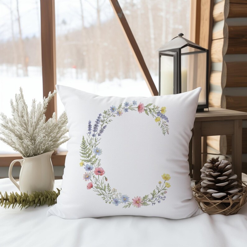 May include: White square pillow with a floral wreath in the shape of the letter "C". The wreath features a variety of colorful flowers and greenery. The pillow is on a white surface, with a window and decorative items in the background.