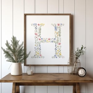 Könnte beinhalten: Gerahmter Blumendruck mit dem Buchstaben "H", der aus bunten Wildblumen besteht. Das Kunstwerk befindet sich vor einem weißen Hintergrund und ist in einem braunen Holzrahmen gerahmt. Der Druck wird auf einem Holztisch ausgestellt.