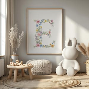 May include: A framed floral print featuring the letter "E" composed of watercolor flowers in pink, blue, and yellow. The artwork is displayed in a light-colored wooden frame. The room also includes a white bunny statue, a small wooden table, and a woven rug.