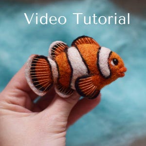 May include: A handmade felt clownfish in orange, white, and black. The fish has detailed fins and a small eye. The text "Video Tutorial" is above the fish. The fish is held in a hand against a blue background.
