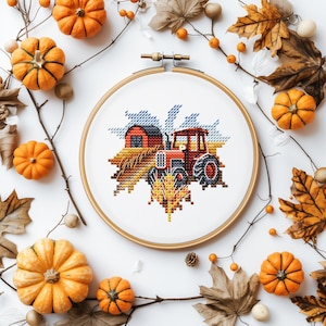 May include: A cross-stitch embroidery hoop featuring a red tractor and barn scene, surrounded by orange pumpkins, fall leaves, and berries. The embroidery is set against a white background, creating a seasonal autumn theme.