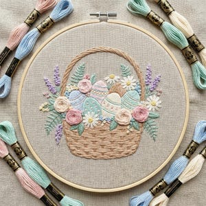 May include: An embroidered Easter basket filled with decorated eggs, flowers, and greenery. The basket is beige and surrounded by a wooden embroidery hoop. Spools of pastel-colored embroidery floss are arranged around the hoop.