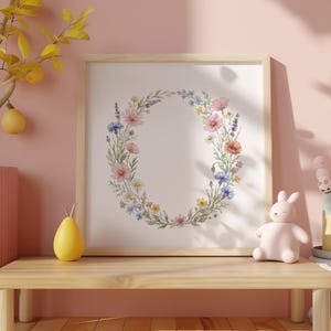 Op de afbeelding: Een ingelijste bloemenkransprint met roze, blauwe en gele bloemen. Het kunstwerk wordt weergegeven op een lichte houten tafel met een roze konijnenbeeldje en een geel peervormig object.