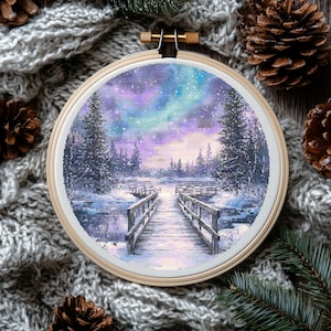 Op de afbeelding: Een borduurwerk met kruissteekjes dat een winterscène voorstelt met een houten brug die naar een besneeuwd bos leidt. De lucht is een levendig paars en blauw met een vleugje roze. De scène is ingelijst in een houten ring.