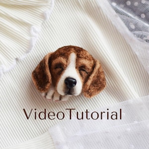 May include: A needle felted beagle puppy with brown and white fur, sleeping with its eyes closed. The puppy is lying on a white fabric background with the text "Video Tutorial" below it.