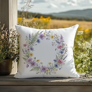 May include: White square pillow with a floral wreath design. The wreath features a variety of flowers in shades of purple, pink, yellow, and blue, with green fern accents. The pillow is placed on a wooden surface with a natural outdoor background.