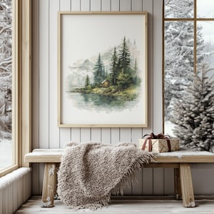 Può includere: Un dipinto ad acquerello incorniciato di una baita sul lago circondata da alberi sempreverdi. L'opera d'arte è esposta su una parete bianca sopra una panca di legno con una coperta beige e un regalo avvolto. La scena è ambientata vicino a una finestra con vista sulla neve.