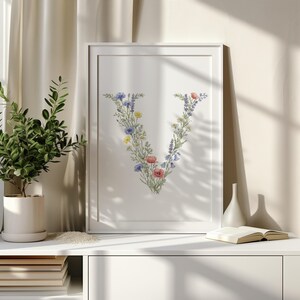 May include: A framed print featuring the letter "V" artfully composed of colorful wildflowers. The floral arrangement includes blue, yellow, and red blooms, set against a white background. The artwork is displayed in a white frame, suitable for home decor.