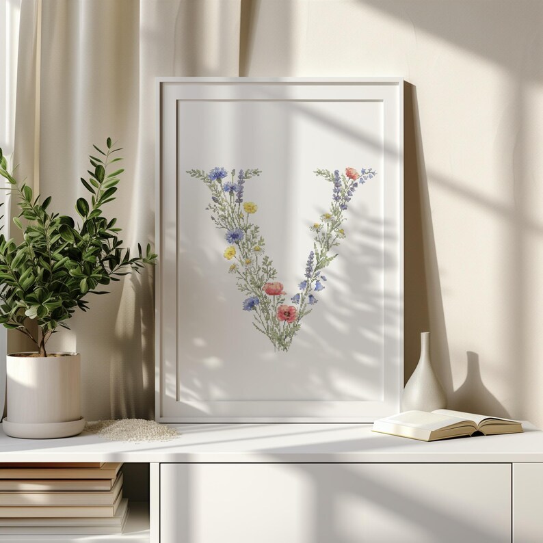 May include: A framed print featuring the letter "V" artfully composed of colorful wildflowers. The floral arrangement includes blue, yellow, and red blooms, set against a white background. The artwork is displayed in a white frame, suitable for home decor.