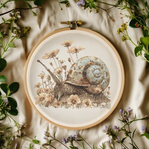 Puede incluir: Un aro de bordado de punto de cruz con un fondo beige que presenta una imagen detallada de un caracol arrastrándose por un campo de flores delicadas. La concha del caracol es de color azul claro y marrón.