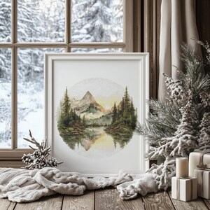 May include: A framed watercolor print of a mountain landscape with a lake, trees, and a sunset. The artwork is in a white frame, displayed on a wooden surface with a cozy, knitted blanket and decorative pine cones. The background shows a snowy winter scene.