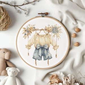 May include: A cross-stitch embroidery hoop with a white background featuring a beige and blue outfit with a bow hanging from a branch with flowers.