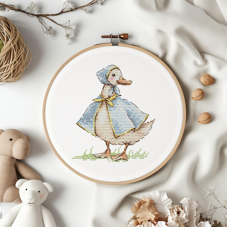Puede incluir: Un bastidor de bordado de punto de cruz con un encantador pato que lleva una capa y un gorro azules con un lazo amarillo. El pato est&aacute; sobre hierba verde, sobre un fondo blanco. El bastidor de madera es visible y el dise&ntilde;o es detallado.
