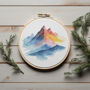 May include: A colorful embroidery hoop with a cross-stitch design of a mountain range with a watercolor effect. The mountains are shades of blue, pink, and yellow.