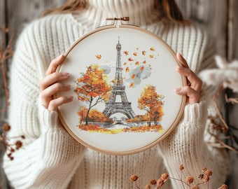 May include: A white embroidery hoop with a cross-stitch design of the Eiffel Tower in Paris, France. The design features the tower in the center with trees and falling leaves on either side. The background is a light blue color.