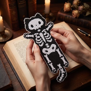May include: A black and white cross-stitch bookmark shaped like a cat skeleton. The design features a pixelated white skull, rib cage, pelvic bone, and tail bones embroidered onto a solid black fabric background with a distinct grid-like texture.