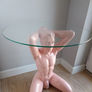 Pode incluir: Uma base de mesa rosa com forma humana com um tampo de vidro transparente. A base é esculpida em uma pose de joelhos com os braços levantados acima da cabeça.