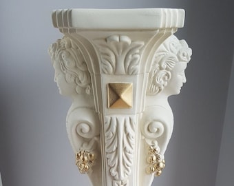 May include: White pedestal with gold accents and ornate carvings. The pedestal has a square base and a round top. It features two cherub-like faces on each side of the top.