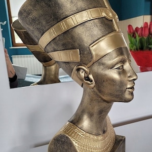 May include: A gold-coloured bust sculpture of an Egyptian queen wearing a headdress with intricate details. The sculpture is on a base with the text "RAZY777" inscribed on it.