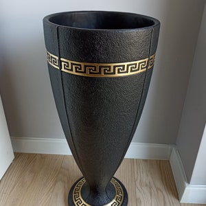 Può includere: Vaso decorativo nero e dorato con un motivo a chiave greca. Il vaso è alto e slanciato con una base larga.