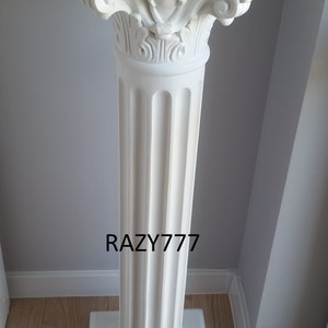 Op de afbeelding: Een wit voetstuk met een vierkante bovenkant en een gegroefde kolom. Het voetstuk heeft sierlijke versieringen aan de boven- en onderkant.