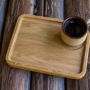 Puede incluir: Una bandeja de madera marrón claro con forma rectangular redondeada. Una taza de cerámica beige con borde marrón oscuro y llena de líquido oscuro se encuentra en la bandeja. La bandeja está colocada sobre un fondo de tablones de madera.