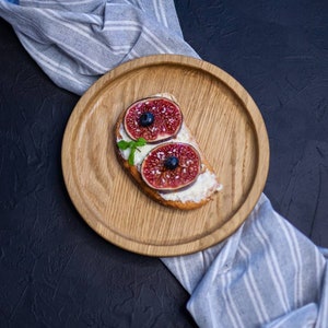 Può includere: Un piatto di legno con una fetta di pane tostato ricoperto di formaggio spalmabile, due metà di fichi freschi e mirtilli. Il piatto è su una superficie nera con un panno a strisce blu e bianco.