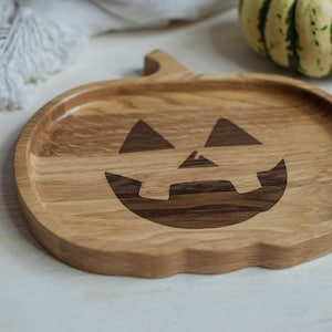 May include: A wooden serving tray shaped like a pumpkin with a carved jack-o'-lantern face. The tray is made of light brown wood with darker brown wood accents.