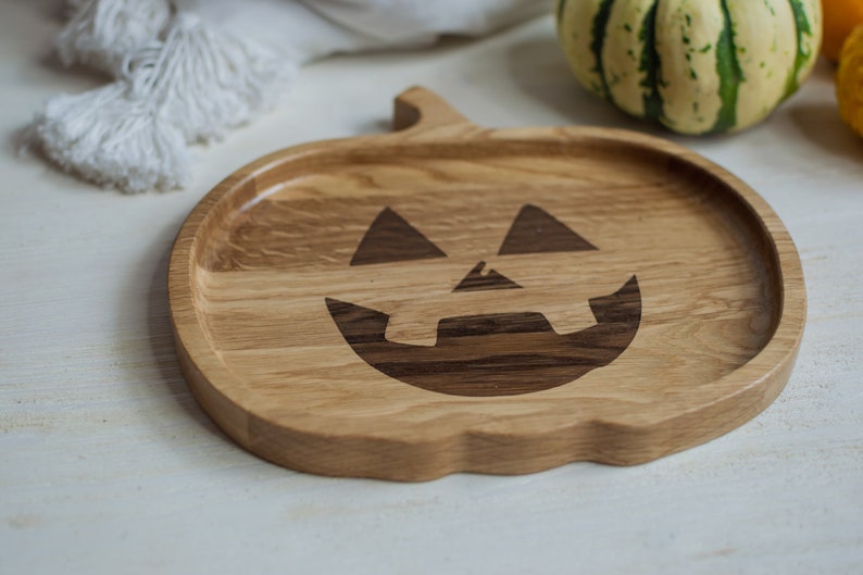 May include: A wooden serving tray shaped like a pumpkin with a carved jack-o'-lantern face. The tray is made of light brown wood with darker brown wood accents.