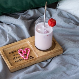 Puede incluir: Una bandeja de madera con una bebida rosa en un vaso con una pajita y una fresa en la parte superior. La bandeja tiene una cita que dice "El ritual del café es un tipo de meditación matutina". Hay dos bastones de caramelo rosas en la bandeja.