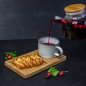 Puede incluir: Una taza de cerámica blanca con una bandeja de madera, un pastel y bayas rojas. La taza se está llenando de té rojo de una tetera de vidrio con una tapa de madera.