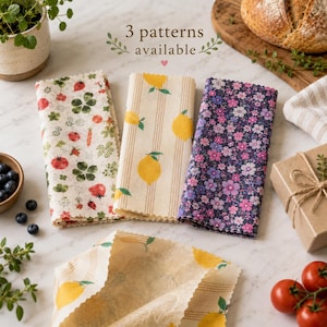 May include: Three rectangular beeswax food wraps featuring distinct prints: one with red strawberries and green leaves, one with yellow lemons on vertical stripes, and one with a dense pattern of small pink and purple flowers on a dark background.